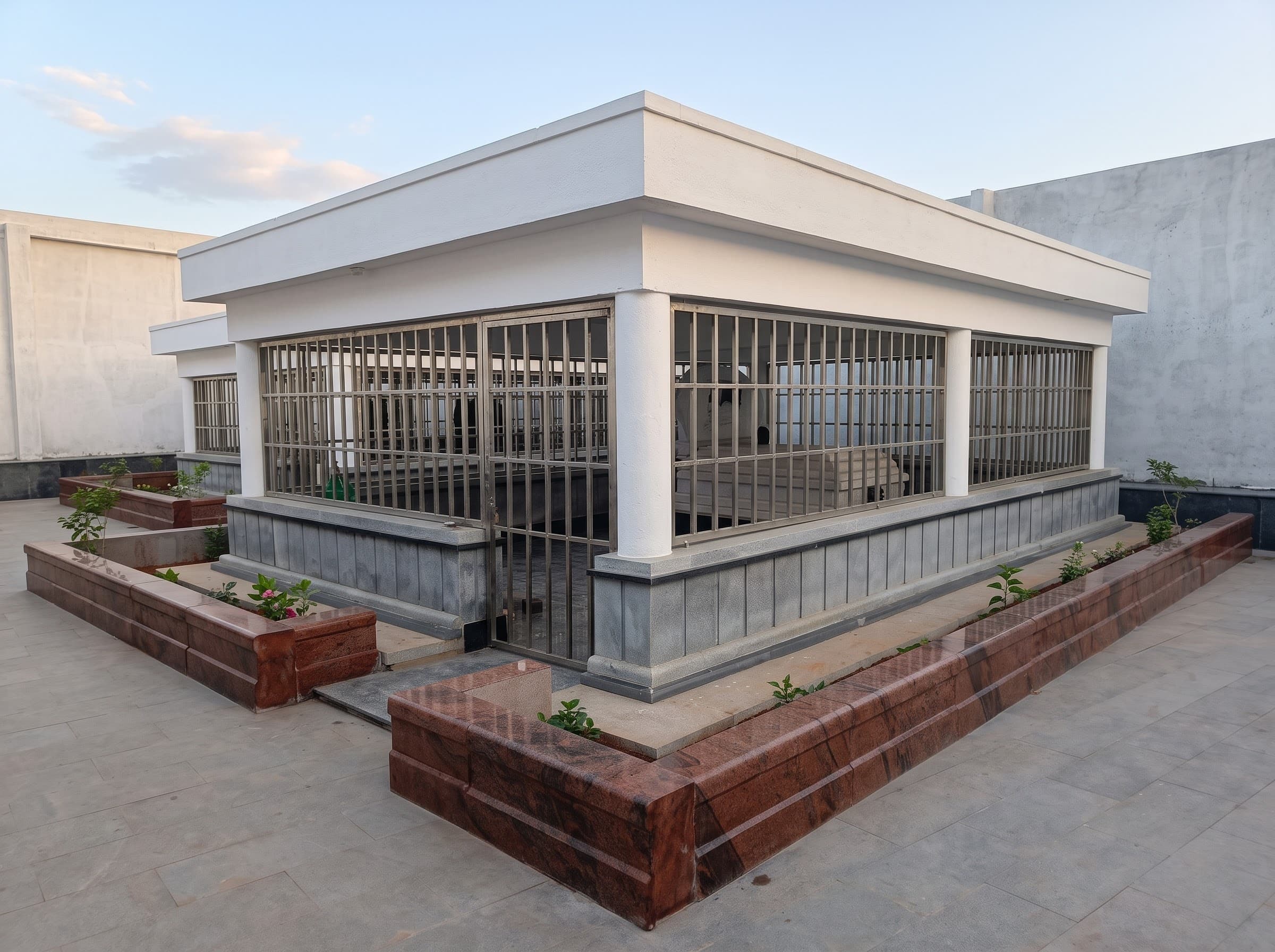 Enclosed granite samadhi structure with grille walls and red granite planters